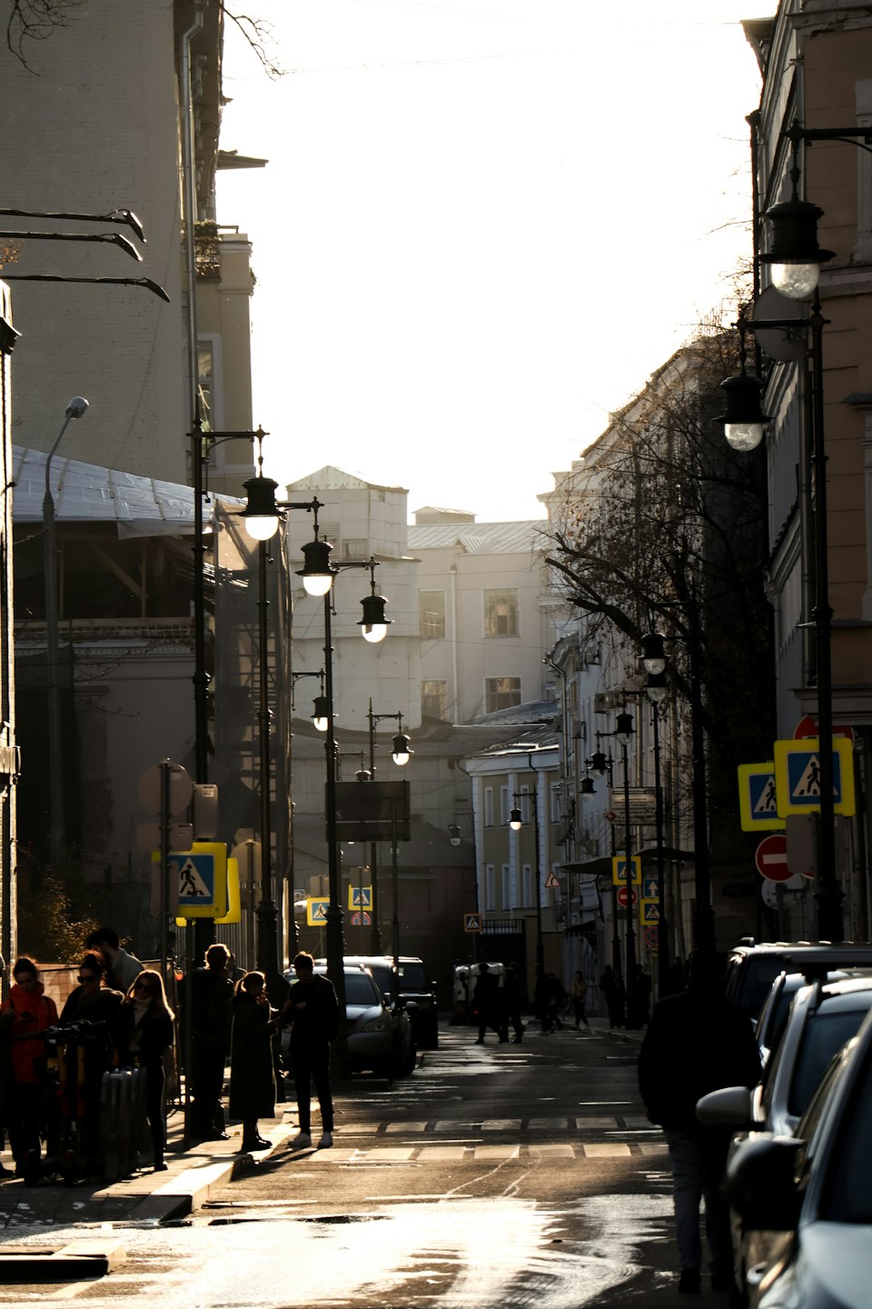 Przechodnie spacerują mokrą ulicą miasta między wysokimi budynkami.